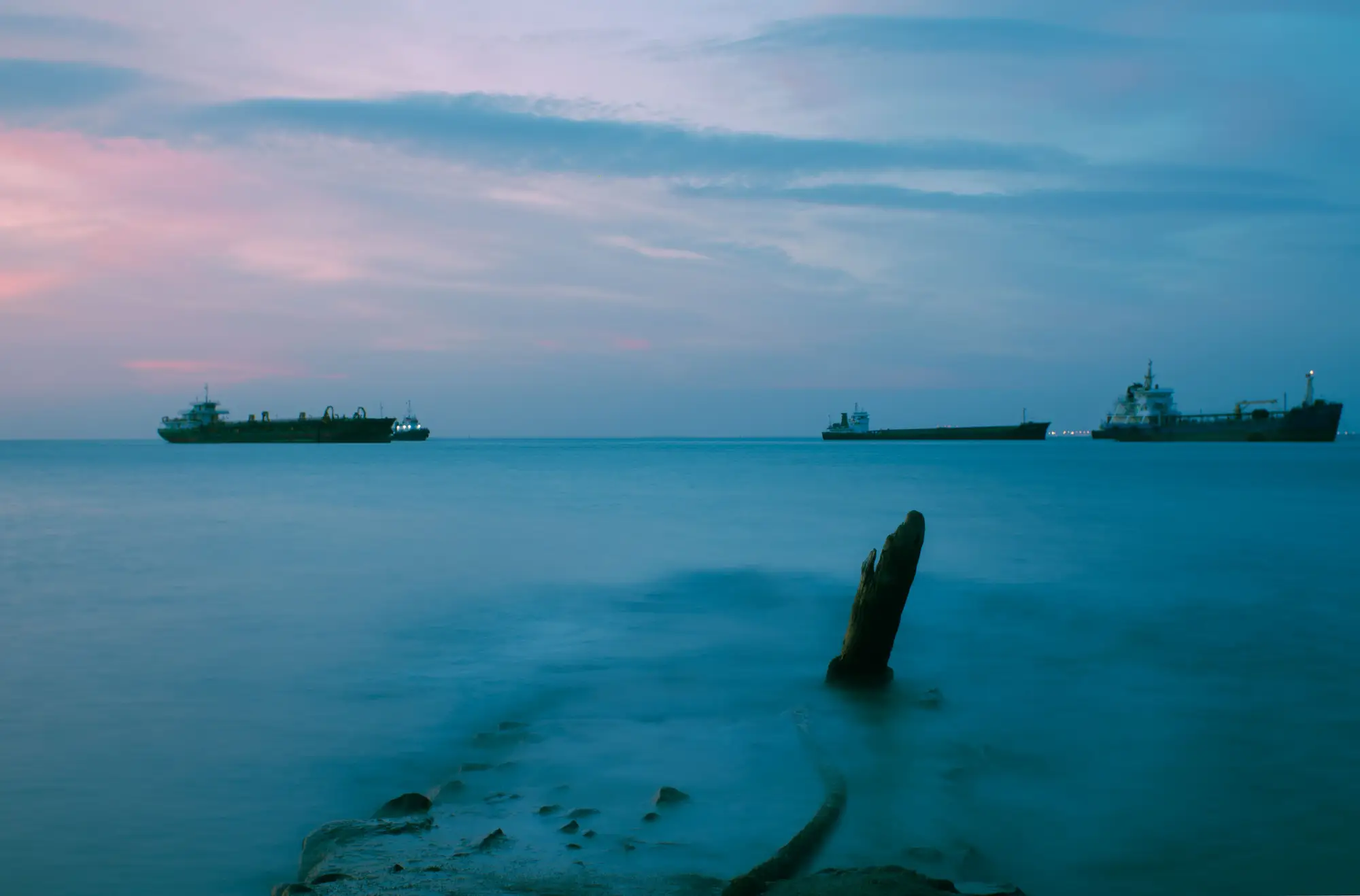 Three cargo ships at anchor on a still sea under a pink and blue evening sky